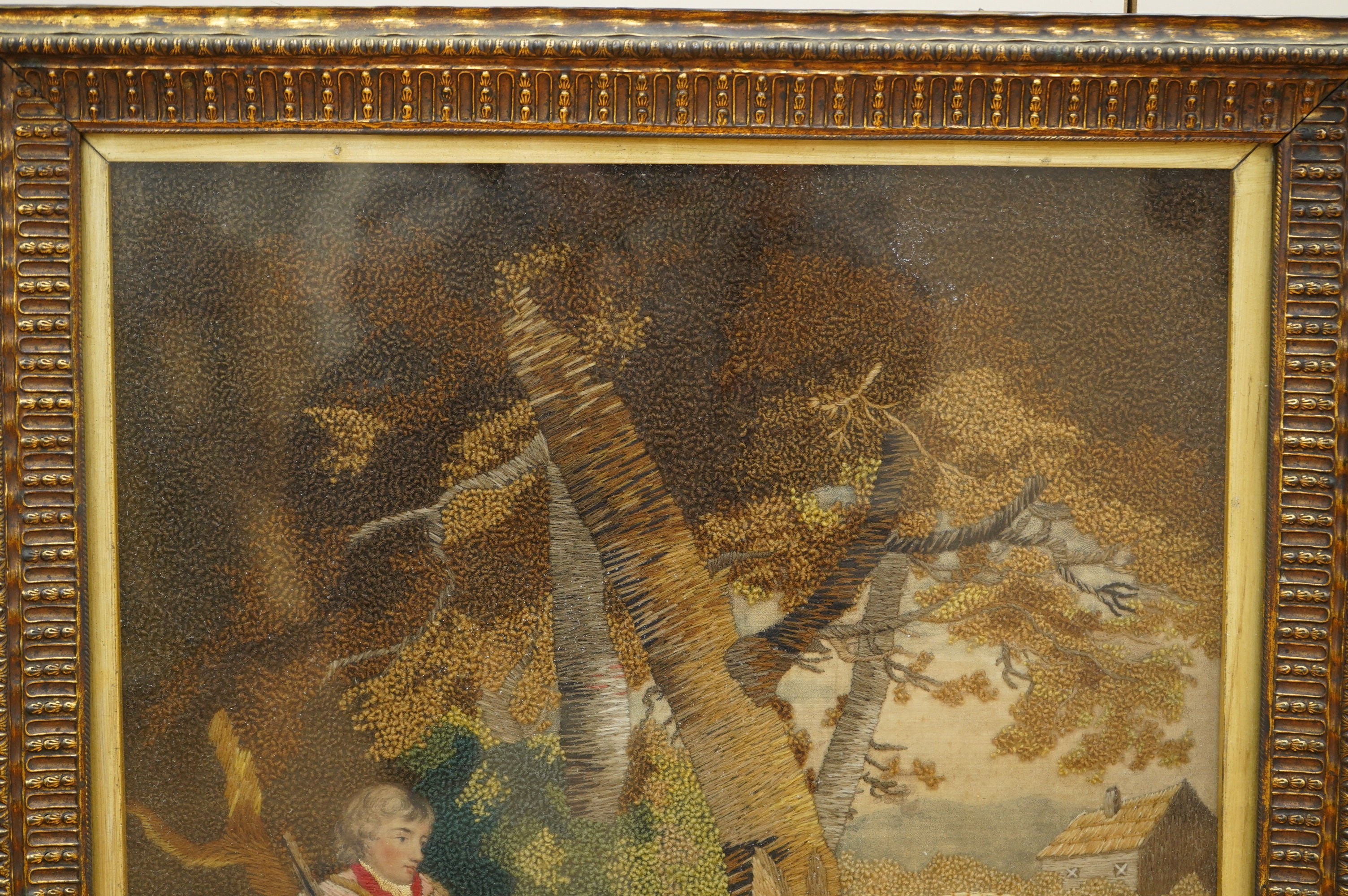 A framed early 19th century wool worked embroidery of huntsman sitting with a rabbit and his three hunting dogs under a tree, by a stream, with a field and cottage in the background, the embroidery worked in fine multi-c